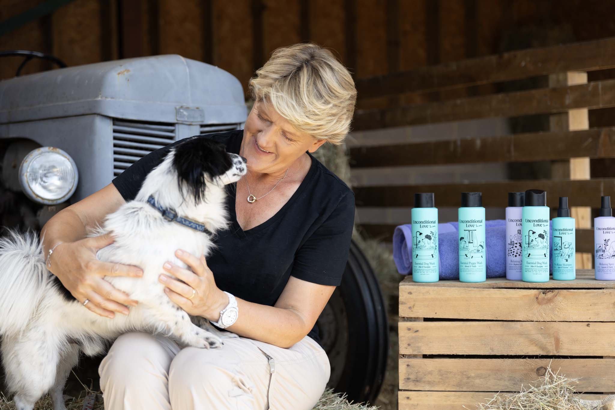 A woman crouches outdoors holding a black-and-white dog, with several pet care bottles arranged on wooden crates beside her. The setting includes a rustic tractor and wooden wall, making this a Lifestyle image.