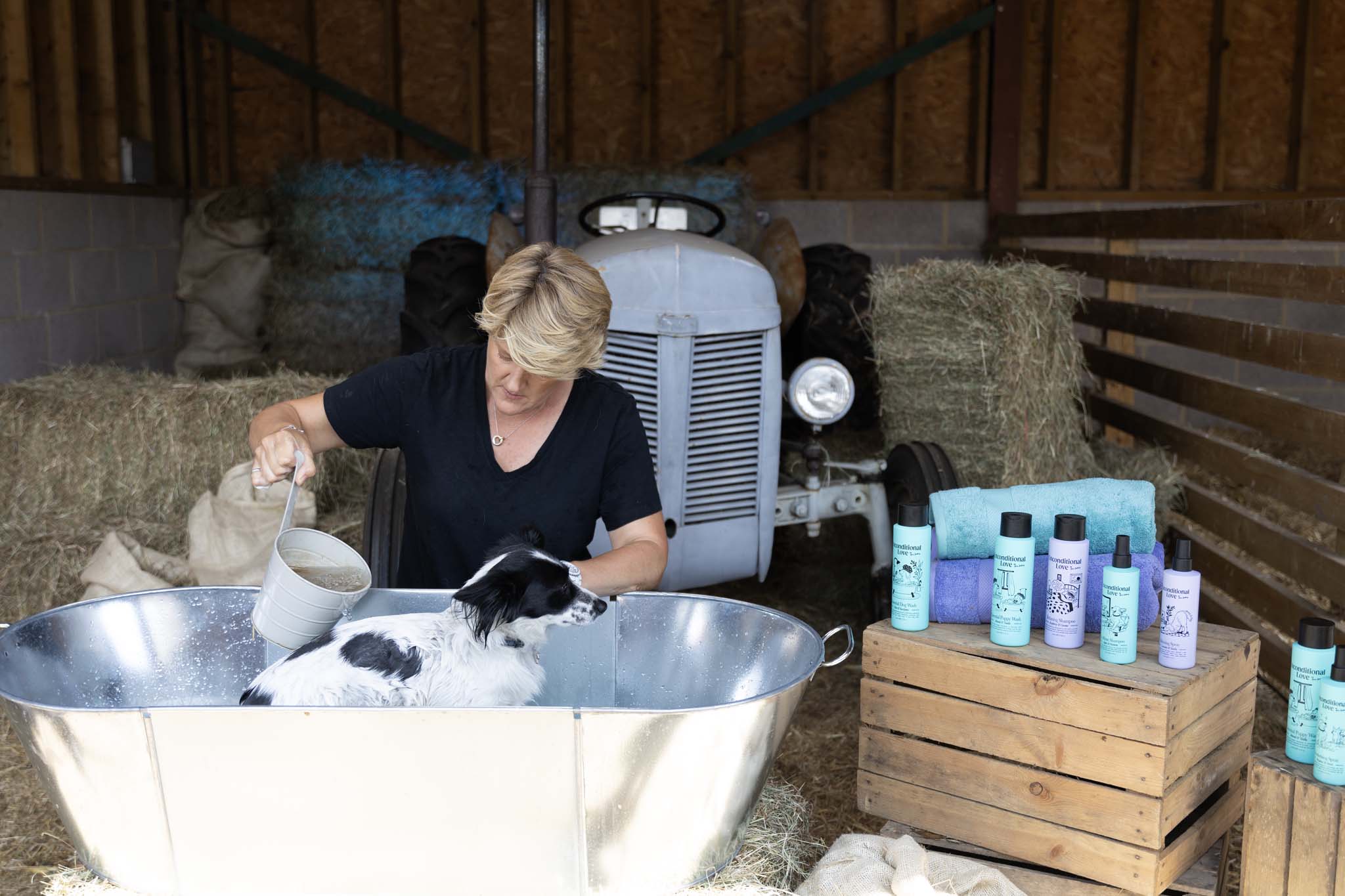 A woman washes a black and white dog in a large metal tub on a farm, with hay bales and a vintage tractor in the background. Several bottles of dog shampoo and conditioner are displayed on a wooden crate to the right.