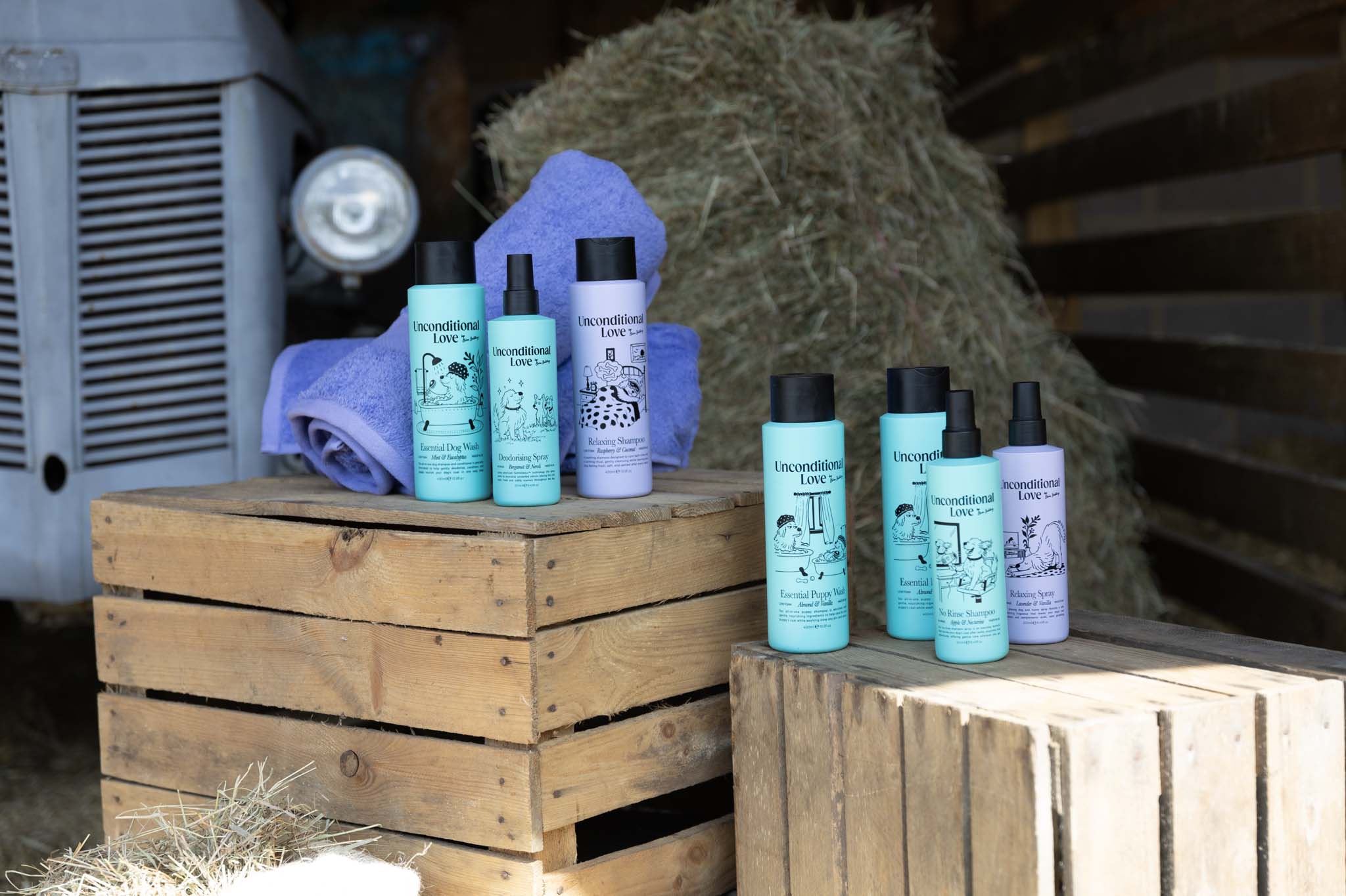 A set of six Unoncditonal Love grooming products, including shampoos and conditioners in turquoise and lavender bottles, displayed on rustic wooden crates with a blue towel and hay in the background.