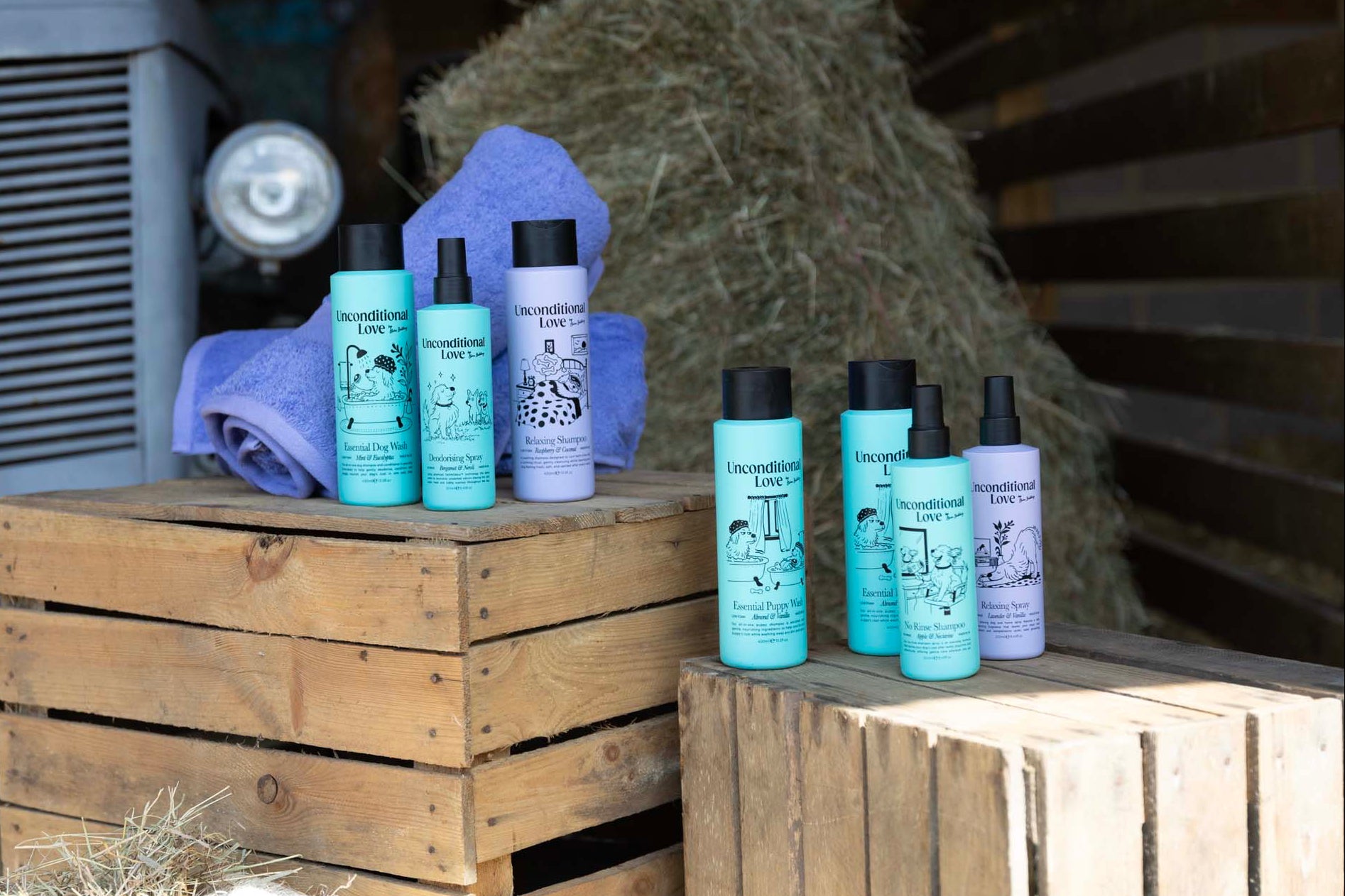 A set of six Unconditional love grooming products, including shampoos and conditioners in turquoise and lavender bottles, displayed on rustic wooden crates with a blue towel and hay in the background.
